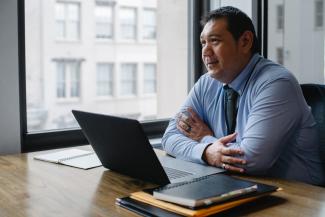 Professional-looking man sitting in an office. There is a laptop and a stack of files in front of him.